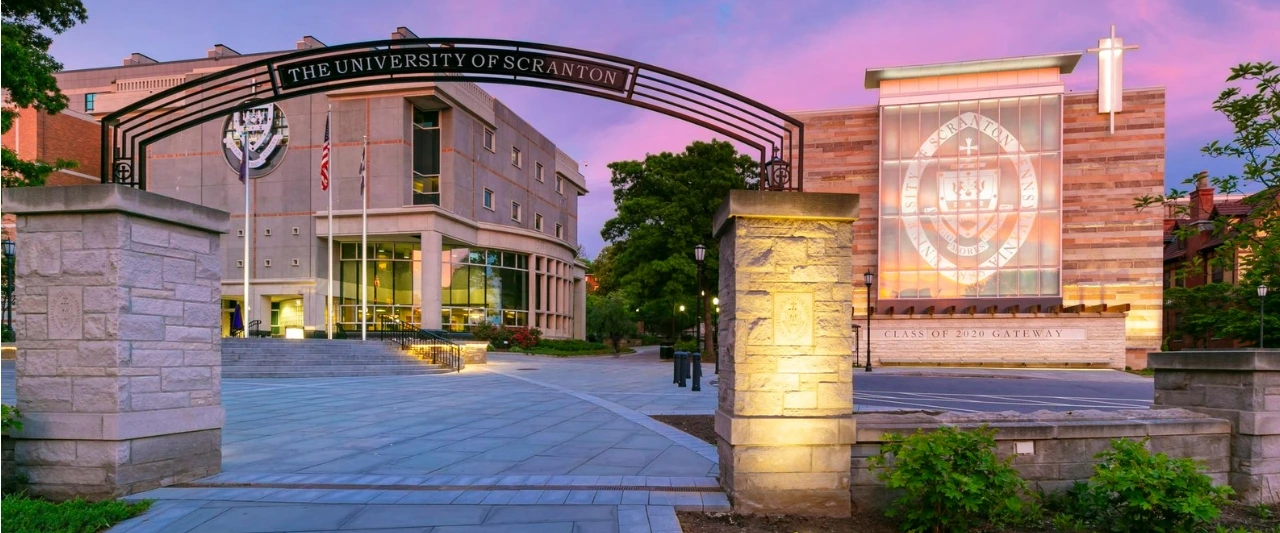 The University of Scranton campus at dusk, featuring a grand stone archway with THE UNIVERSITY OF SCRANTON prominently displayed across the top