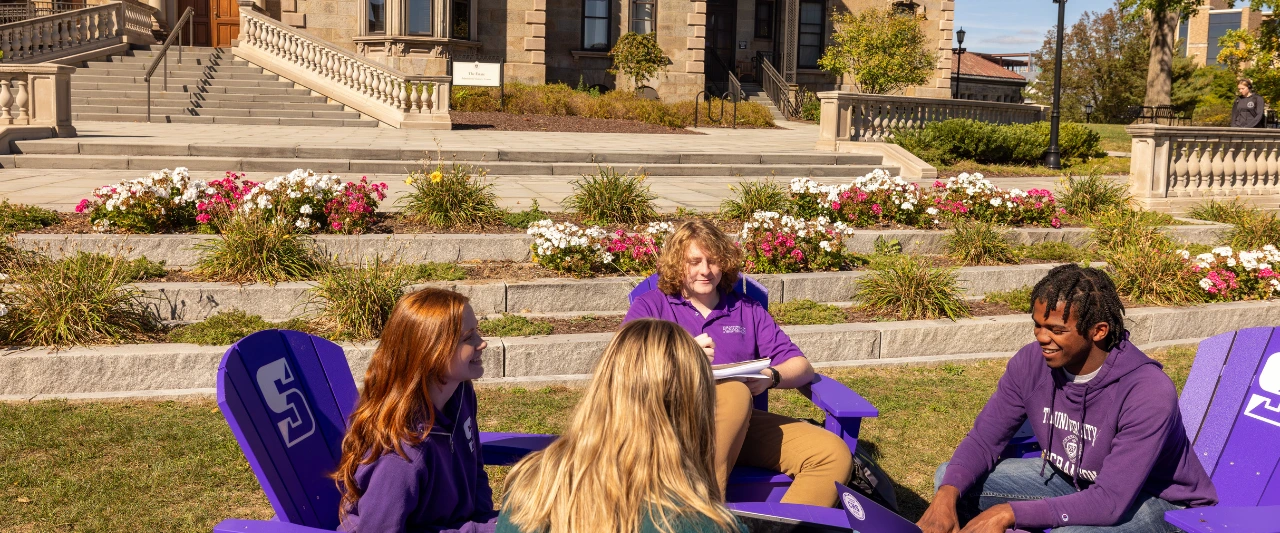 Five Scranton students sit in purple Adirondack chairs on a grassy area in front of The Estate campus building