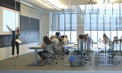 A professor stands at the front of a modern classroom pointing to a presentation slide, while students seated at desks take notes and listen attentively.
