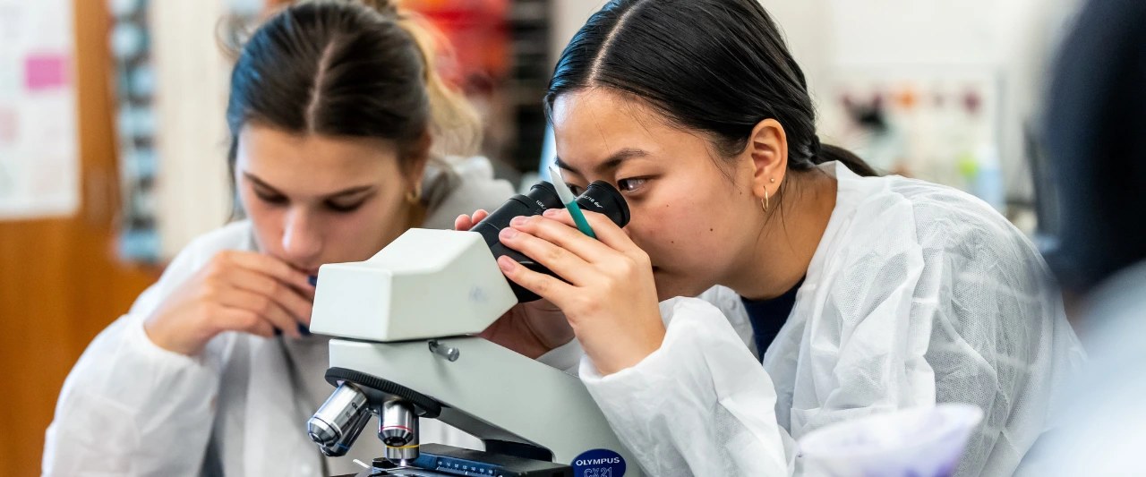 Two Scranton students in lab coats working with a microscope in a classroom