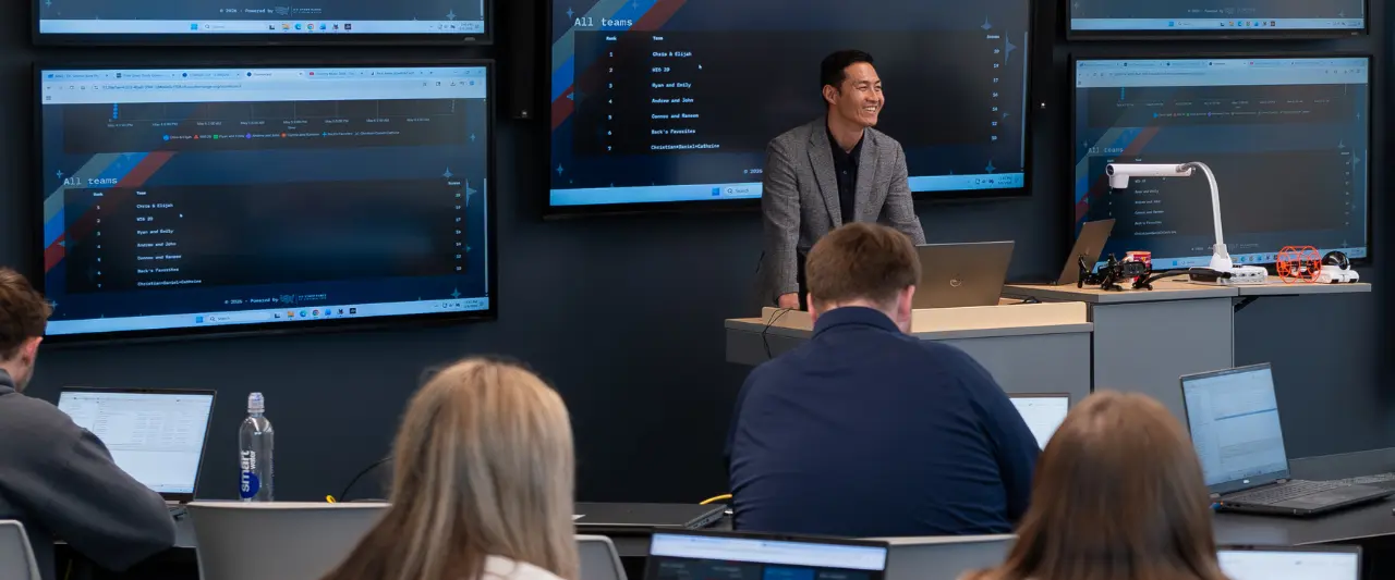A smiling Scranton professor stands at a podium in a modern lab with large screens showing a team leaderboard as students sit at desks using laptops