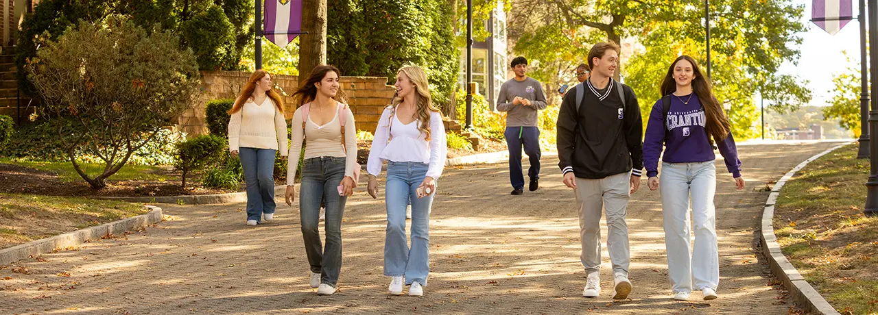 A group of University of Scranton students walk and talk together along a tree-lined campus path on a sunny day