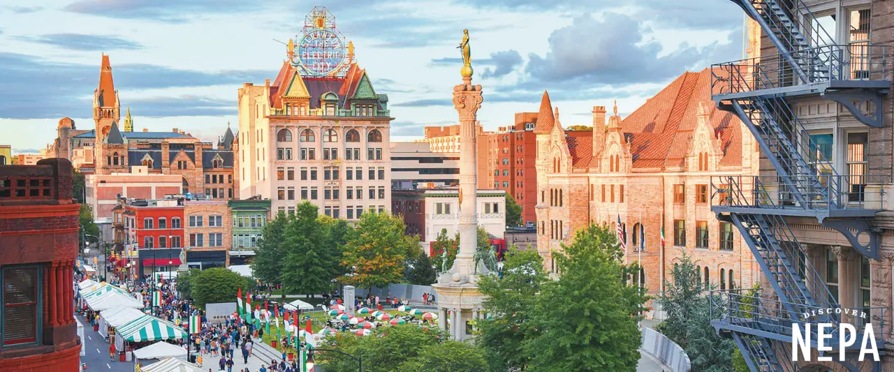 Downtown Scranton scene during La Festa Italian festival with crowds, vendor tents, a central tall monument, historic buildings, and the Scranton Electric City sign