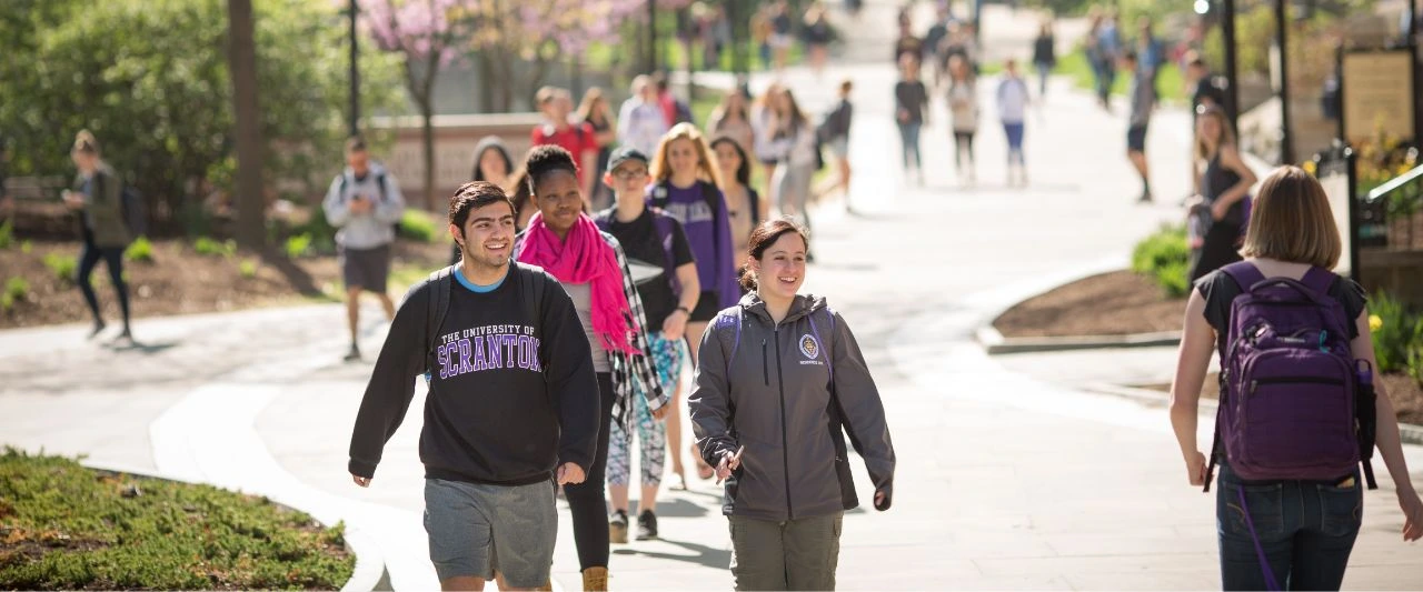 A group of Scranton college students walks along a campus pathway on a sunny spring day
