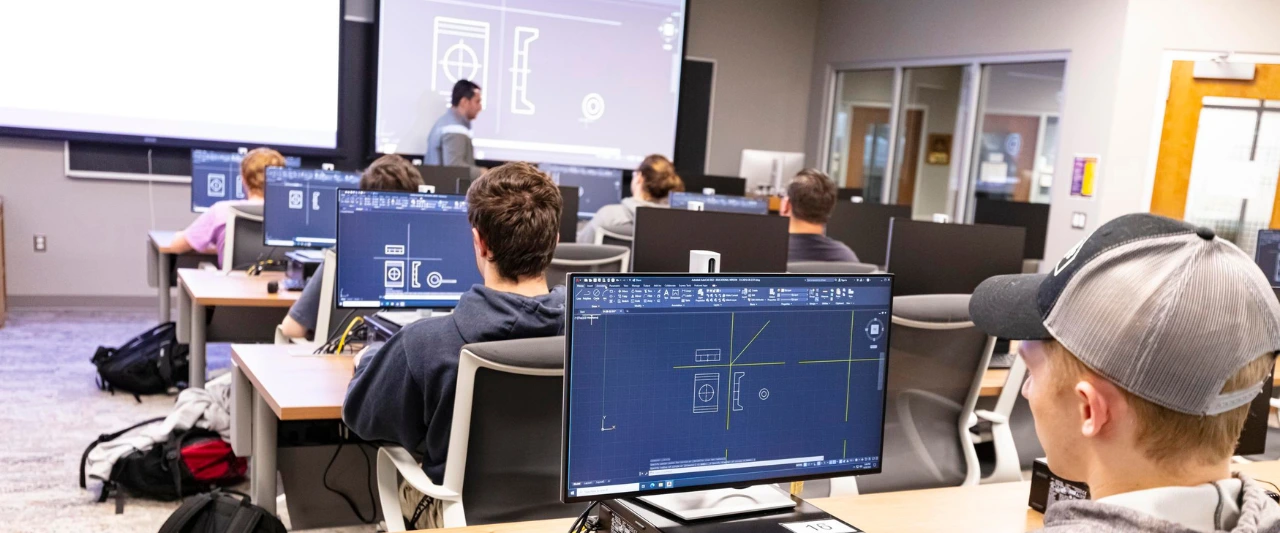 Engineering students during class work at individual computer workstations and follow the professor's instructions to replicate the CAD technical drawings and geometric shapes displayed on the large projection screen in front of the classroom