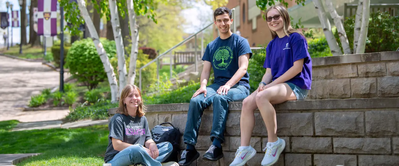 Three students in University of Scranton apparel smiling outside, with two of them sitting on a low stone wall while the third is sitting comfortably on the grass below